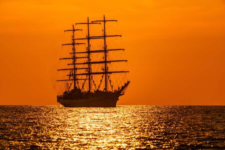 A red burning sunset with the silhouette of sailing ship. Sailors set sails on masts of ship. White sailing vessel floating in the sea. Copy space.の写真素材