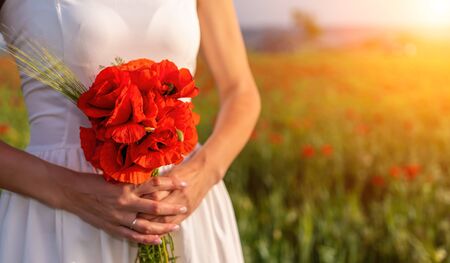 Bride in a white dress holding a bouquet of poppy flowers, warm sunset time on the background of the red poppy field. Copy space. The concept of calmness, silence and unity with nature.の写真素材