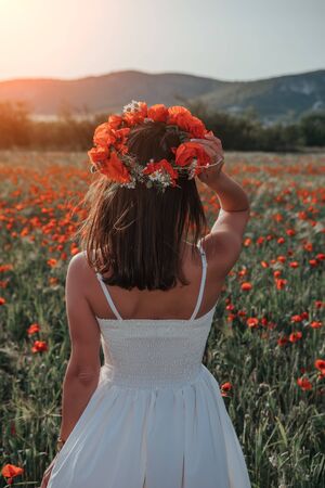 Bride in a white dress in a wreath of red poppies, warm sunset time on the background of the big red poppy field. Copy space. The concept of calmness, silence and unity with nature.の写真素材