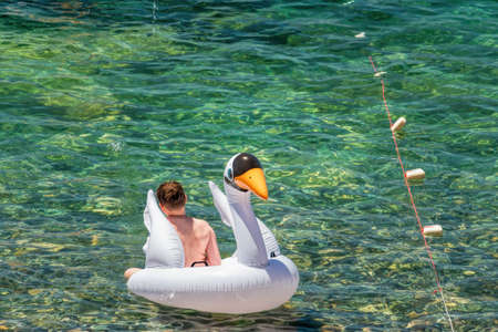 Summer vacation boy relaxing in sea on white inflatable swan swimming pool air mattress bed floating in turquoise water background. Suntan at tropical beachの写真素材
