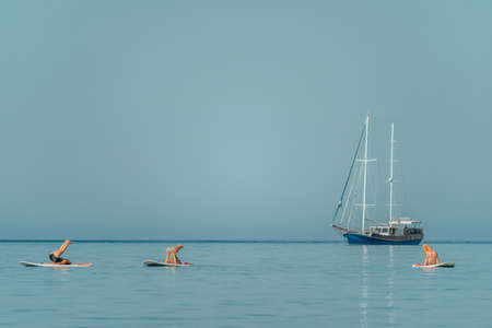 Group of young womens in swimsuits doing yoga on sup board in calm sea, early morning. Balanced pose...