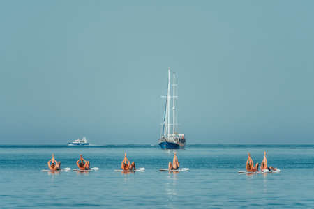 Group of young womens in swimsuits doing yoga on sup board in calm sea, early morning. Balanced pose...