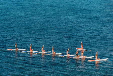 Group of young womens in swimsuits doing yoga on sup board in calm sea, early morning. Balanced pose - concept of healthy life and natural balance between body and mental development.の写真素材