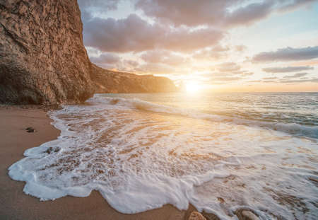 Bright sea sunset. The waves crash into the rock, lit by the warm sunset, sand and pebbles, volcanic basalt as in Iceland. Sea wave breaks into splashes and white foam. Never-ending beauty of natureの写真素材