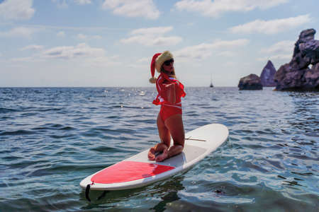 Happy young attractive brunette woman in red swimsuit and Santa hat, swimming on sup board in the...