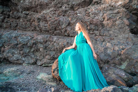 Side view a Young beautiful sensual woman in a mint long dress posing on a rock high above the sea during sunrise. Girl on the nature on blue sky background. Fashion photoの写真素材