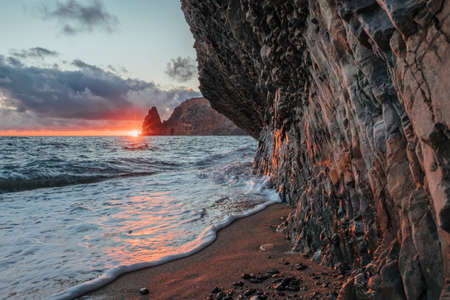 Bright sea sunset. The waves crash into the rock, lit by the warm sunset, sand and pebbles, volcanic basalt as in Iceland. Sea wave breaks into splashes and white foam. Never-ending beauty of natureの写真素材