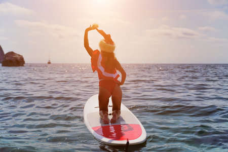 Happy young attractive brunette woman in red swimsuit and Santa hat, swimming on sup board in the...