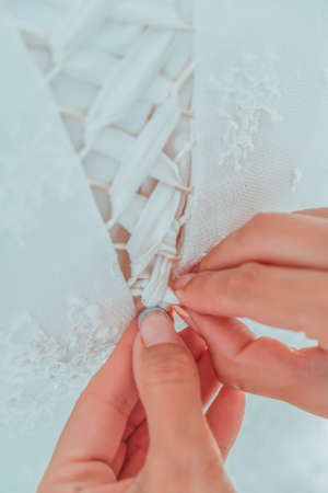 Morning preparation of the bride. Closeup of bridesmaid tying bow on brides wedding dress. White ribbon bow on the back of wedding dress. Bride in white wedding dress with lace standing in the roomの写真素材