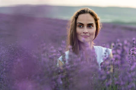 Close up portrait of happy young brunette woman in white dress on blooming fragrant lavender fields with endless rows. Warm sunset light. Bushes of lavender purple aromatic flowers on lavender fields.の写真素材