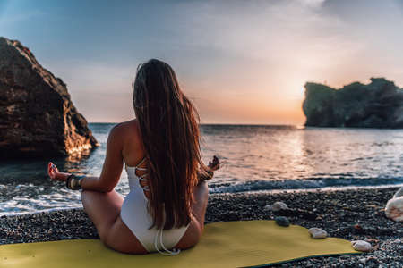 Young woman with long hair in white swimsuit and boho style braclets practicing outdoors on yoga mat by the sea on a sunset. Womens yoga fitness routine. Healthy lifestyle, harmony and meditationの写真素材