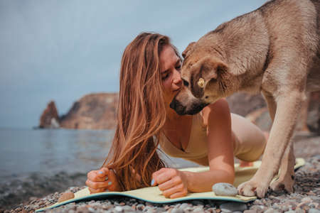 Young woman in beige sportswear with long hair practicing stretching outdoors on yoga mat by the sea on a sunny day. Womens yoga fitness pilates routine. Healthy lifestyle and meditation conceptの写真素材