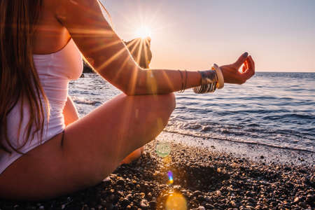 Young woman with long hair in white swimsuit and boho style braclets practicing outdoors on yoga mat by the sea on a sunset. Womens yoga fitness routine. Healthy lifestyle, harmony and meditationの写真素材