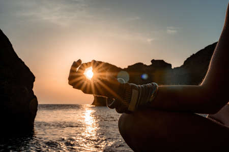 Young woman with long hair in white swimsuit and boho style braclets practicing outdoors on yoga mat by the sea on a sunset. Womens yoga fitness routine. Healthy lifestyle, harmony and meditationの写真素材