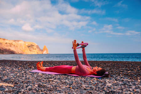Young woman with black hair, fitness instructor in pink sports leggings and tops, doing pilates on yoga mat with magic pilates ring by the sea on the beach. Female fitness daily yoga concept.の写真素材