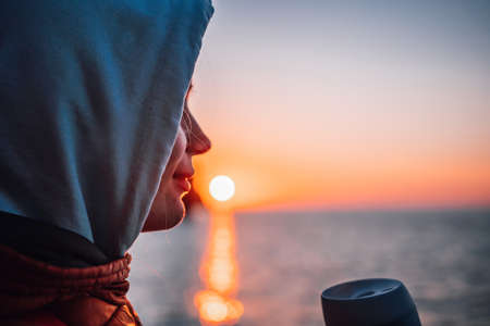 A young tourist Woman holding coffee tumbler cup while sitting outdoor and enjoying sunrise over sea...