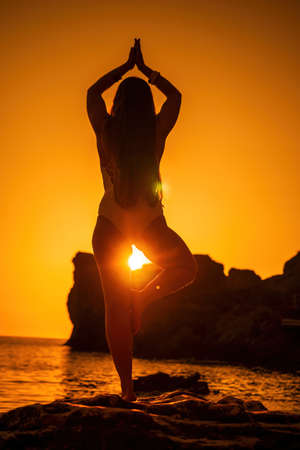 Young woman with long hair in white swimsuit and boho style braclets practicing outdoors on yoga mat by the sea on a sunset. Womens yoga fitness routine. Healthy lifestyle, harmony and meditationの写真素材