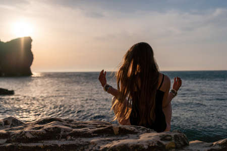 Young woman with long hair in black swimsuit and boho style braclets practicing outdoors on yoga mat by the sea on a sunset. Womens yoga fitness routine. Healthy lifestyle, harmony and meditationの写真素材