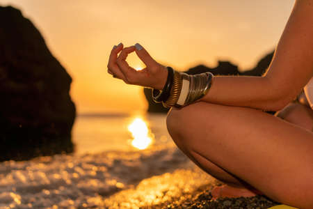 Young woman with long hair in white swimsuit and boho style braclets practicing outdoors on yoga mat by the sea on a sunset. Womens yoga fitness routine. Healthy lifestyle, harmony and meditationの写真素材