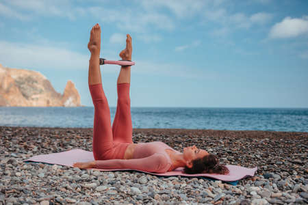 Young woman with black hair, fitness instructor in pink sports leggings and tops, doing pilates on yoga mat with magic pilates ring by the sea on the beach. Female fitness daily yoga concept.の写真素材