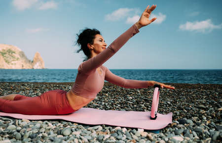 Young woman with black hair, fitness instructor in pink sports leggings and tops, doing pilates on yoga mat with magic pilates ring by the sea on the beach. Female fitness daily yoga concept.の写真素材