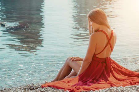 Side view a Young beautiful sensual woman in a red long dress posing on the near the sea during sunrise. Summer fun, holidays travel. Fashion photoの写真素材