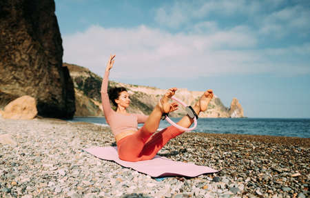 Young woman with black hair, fitness instructor in pink sports leggings and tops, doing pilates on yoga mat with magic pilates ring by the sea on the beach. Female fitness daily yoga concept.の写真素材