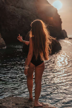 Young woman with long hair in black swimsuit and boho style braclets practicing outdoors on yoga mat by the sea on a sunset. Womens yoga fitness routine. Healthy lifestyle, harmony and meditationの写真素材