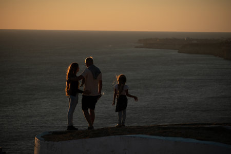 Silhouette of a young romantic couple with a child enjoying the evening on a cliff above the sea...