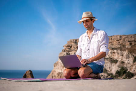 A digital nomad, a man in a hat, a businessman with a laptop does yoga on the rocks by the sea at...
