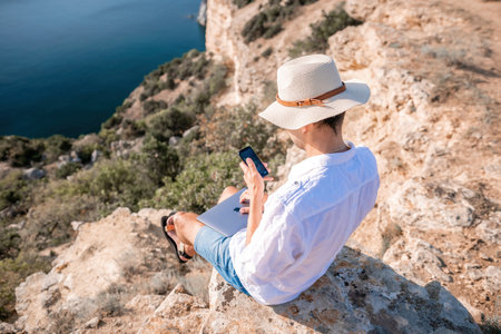 Digital nomad, man in the hat, a businessman with a laptop sits on the rocks by the sea during sunset, makes a business transaction online from a distance. Remote work on vacation.の写真素材