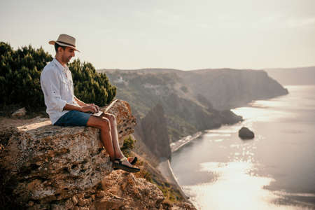 Digital nomad, man in the hat, a businessman with a laptop sits on the rocks by the sea during sunset, makes a business transaction online from a distance. Remote work on vacation.の写真素材