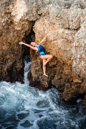 A young beautiful tanned woman with long hair in blue bikini sitting at rocks near blue green clear ocean sea water. Woman relax on sunset at sea, enjoy holidays and weekend vacation in summer time.の写真素材