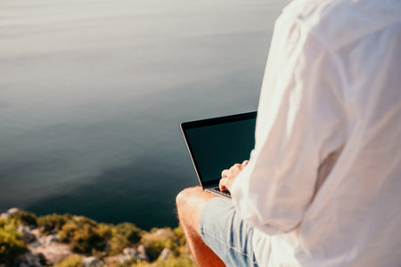 Digital nomad, man in the hat, a businessman with a laptop sits on the rocks by the sea during sunset, makes a business transaction online from a distance. Remote work on vacation.の写真素材