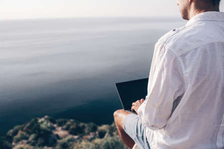Digital nomad, man in the hat, a businessman with a laptop sits on the rocks by the sea during sunset, makes a business transaction online from a distance. Remote work on vacation.の写真素材
