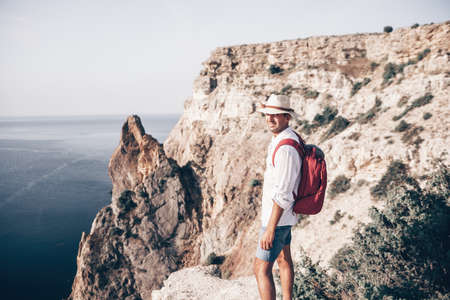 Digital nomad, man in the hat, a businessman with a laptop sits on the rocks by the sea during sunset, makes a business transaction online from a distance. Remote work on vacation.の写真素材