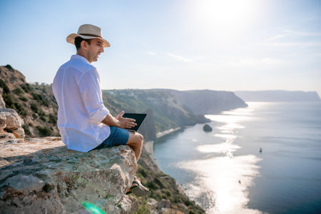 Digital nomad, man in the hat, a businessman with a laptop sits on the rocks by the sea during sunset, makes a business transaction online from a distance. Remote work on vacation.の写真素材