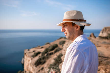 Digital nomad, man in the hat, a businessman with a laptop sits on the rocks by the sea during sunset, makes a business transaction online from a distance. Remote work on vacation.の写真素材