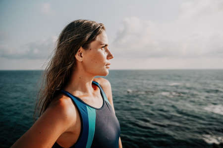 A young beautiful tanned woman with long hair in blue bikini stands at rock near blue green clear ocean sea water. Woman relax on sunset at sea, enjoy holidays and weekend vacation in summer time.の写真素材