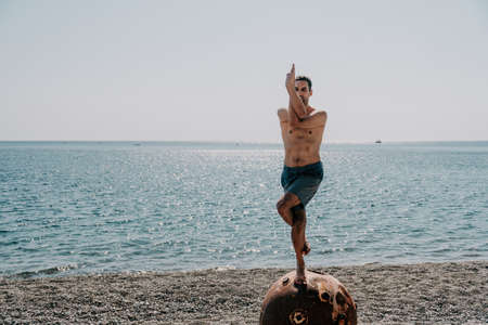 Man doing yoga exercise outdoors standing on old rusty floating marine mine on the beach with rocky shore and sea background. Healthy lifestyle, pollution, nature protection, war and peace concept.の写真素材