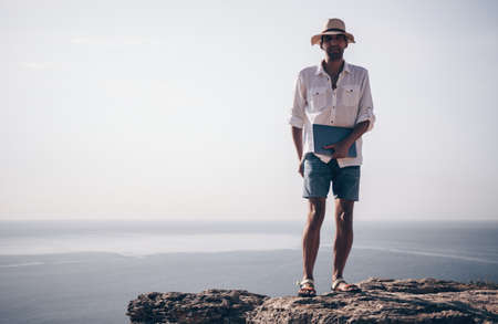 Digital nomad, man in the hat, a businessman with a laptop sits on the rocks by the sea during sunset, makes a business transaction online from a distance. Remote work on vacation.の写真素材