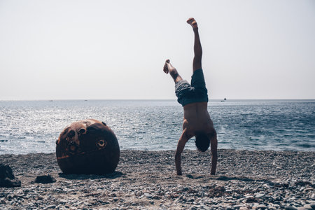 Man doing yoga exercise outdoors standing on old rusty floating marine mine on the beach with rocky...