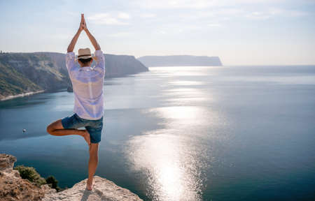 A digital nomad, a man in a hat, a businessman does yoga on the rocks by the sea at sunset time, does a business operation online from a distance. Remote work on vacation.の写真素材