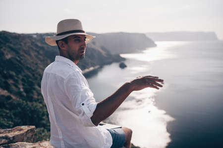 Digital nomad, man in the hat, a businessman with a laptop sits on the rocks by the sea during sunset, makes a business transaction online from a distance. Remote work on vacation.の写真素材