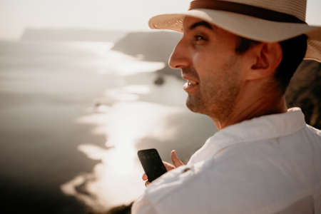 Digital nomad, man in the hat, a businessman with a laptop sits on the rocks by the sea during sunset, makes a business transaction online from a distance. Remote work on vacation.の写真素材