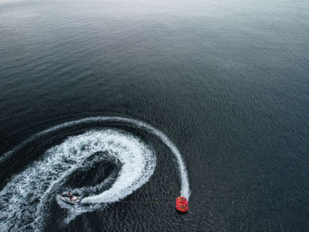 Happy people are going to swim on an air mattress behind a jet ski. Tourists ride the inflatable watercraft boat. Sea attraction. Jet ski skating people on a large Inflatable raft. Aerial copter view.の写真素材