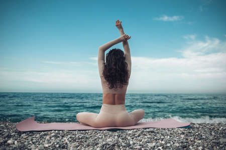 Young woman with black hair, fitness instructor in leggings and tops doing stretching and pilates on yoga mat near the sea. Female fitness yoga routine concept Healthy lifestyle harmony and meditationの写真素材