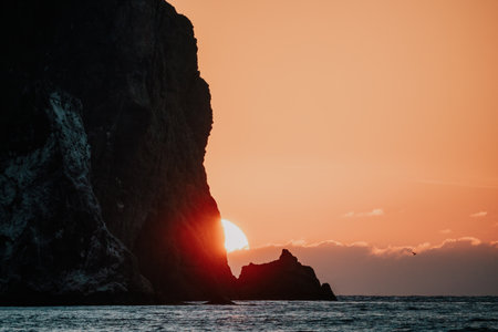 A red burning sunset over the sea with rocky volcanic cliff. Abstract nature summer or spring ocean sea background. Small waves on golden warm water surface with bokeh lights from sun.の写真素材
