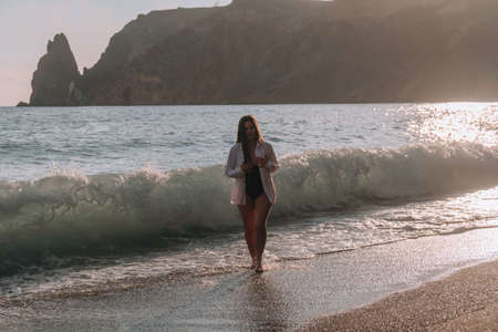 Selective focus. Happy carefree sensual woman with long hair in black swimwear posing at sunset beach. Silhouette of young beautiful playful positive woman outdoor. Summer vacation and trip conceptの写真素材