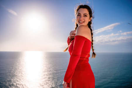 Closeup portrait of sensual young brunette woman in red dress, happily dancing outdoors isolated on blurry sea background with natural bokeh in soft warm sunset backlight. Selective focus.の写真素材
